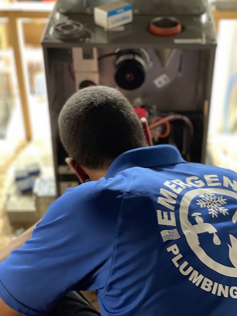 A technician in a blue company shirt marked, "Emergency Plumbing", leans into a heater during maintenance.