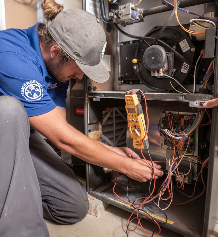 A technician wearing a company shirt is checking the electrical panel of a furnace.