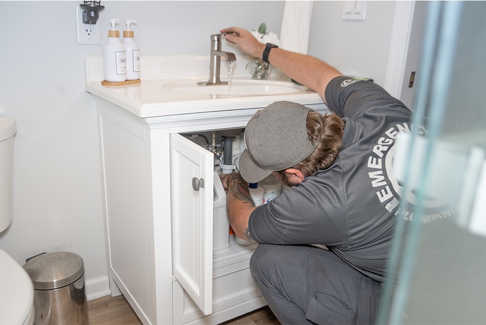 A plumber kneels and examines pipes under a bathroom sink, wearing a company shirt labeled "EMERGENCY PLUMBING".