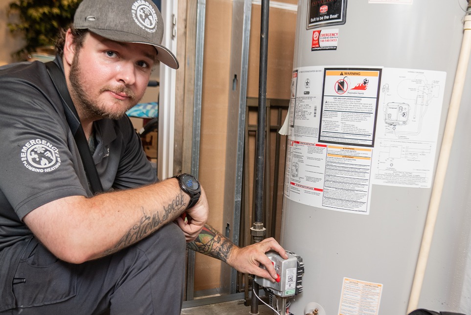 A plumber for Emergency Plumbing Heating & Air kneels to inspect a water heater. 