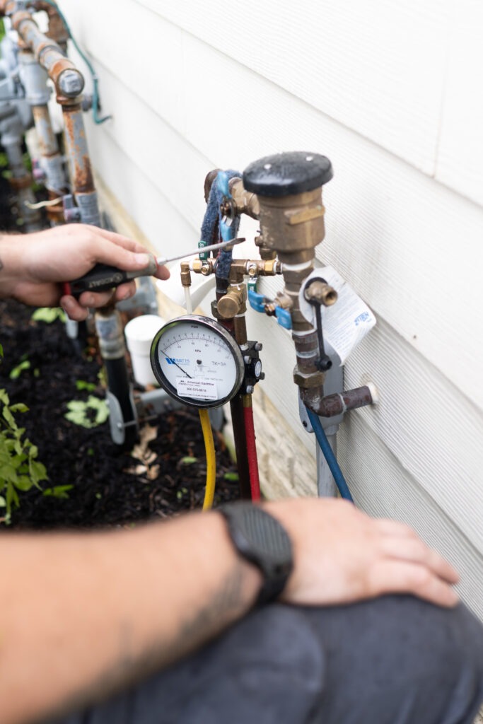 A technician repairs a backflow prevention device mounted on a building exterior with a pressure gauge attached.