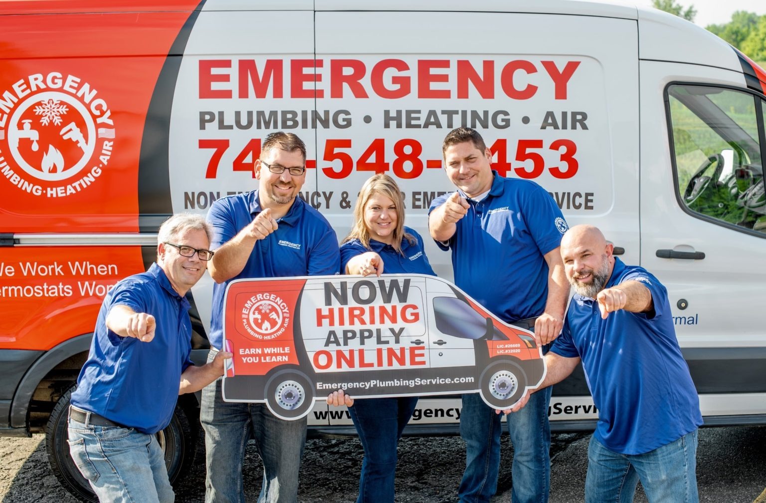 Five staff members from Emergency Plumbing, Heating, and Air point at the camera while holding a sign.