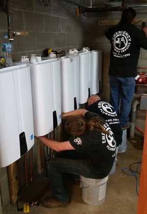 Three workers wearing "Emergency Plumbing Service" shirts install several white water heaters.