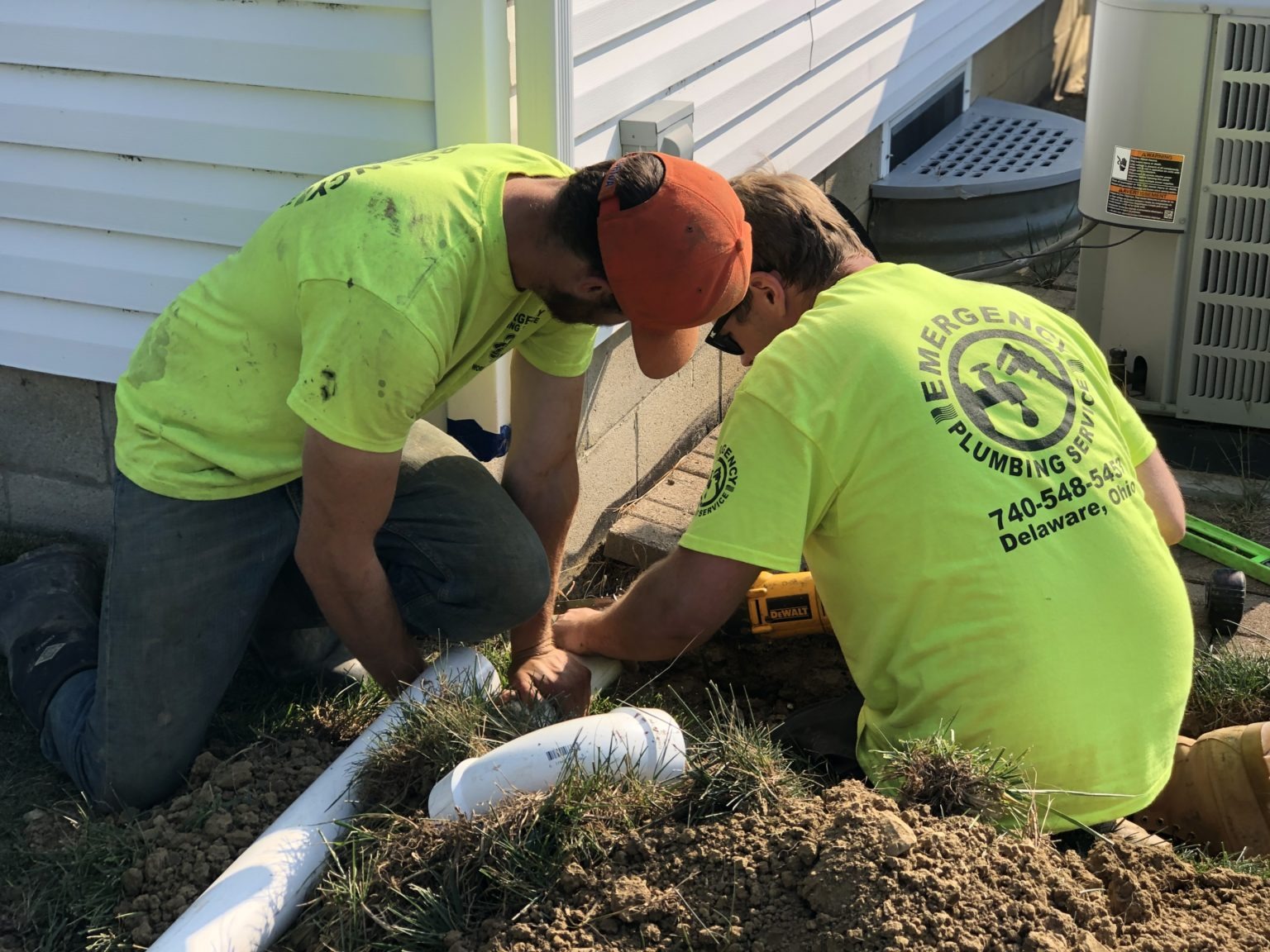 Two plumbers in bright yellow shirts work on white pipes outside a building.