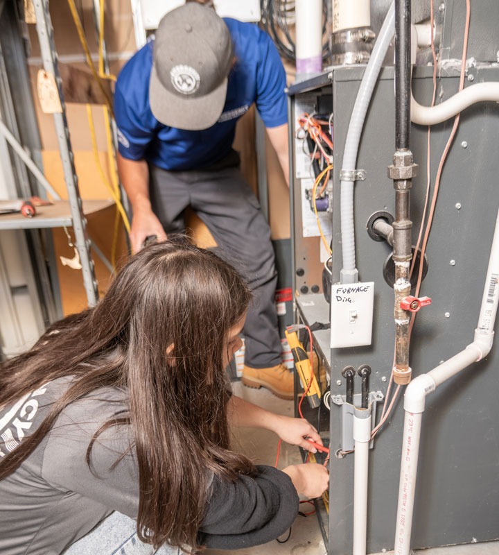 Two technicians work to repair an HVAC system, with the text &quot;FURNACE DNG&quot; visible on a nearby switch.