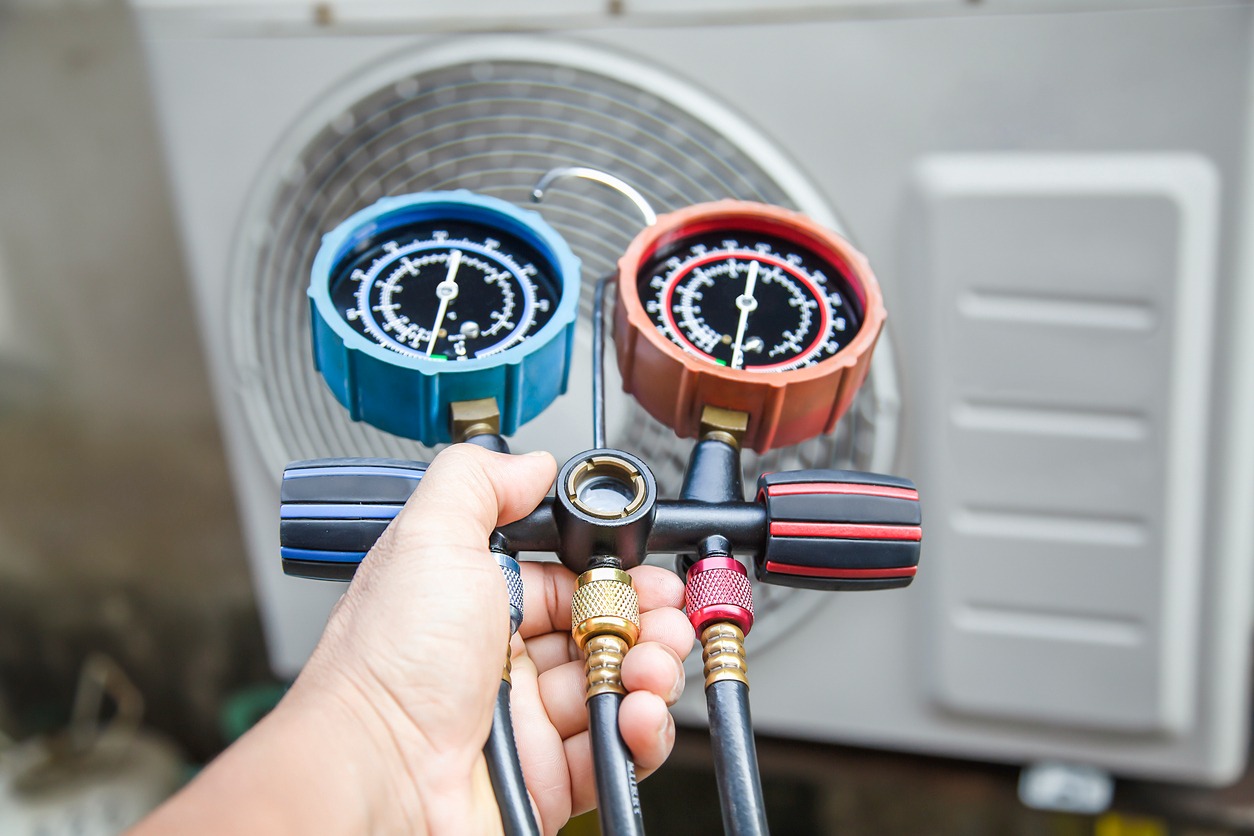 An HVAC technician holds a gauge manifold, likely checking the pressure in an AC unit.