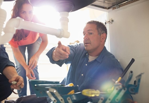 A plumber points to the plumbing under a sink, explaining it to a homeowner.