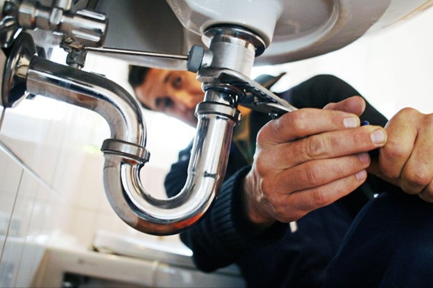 A plumber, under a sink, is fixing a pipe with a wrench. 