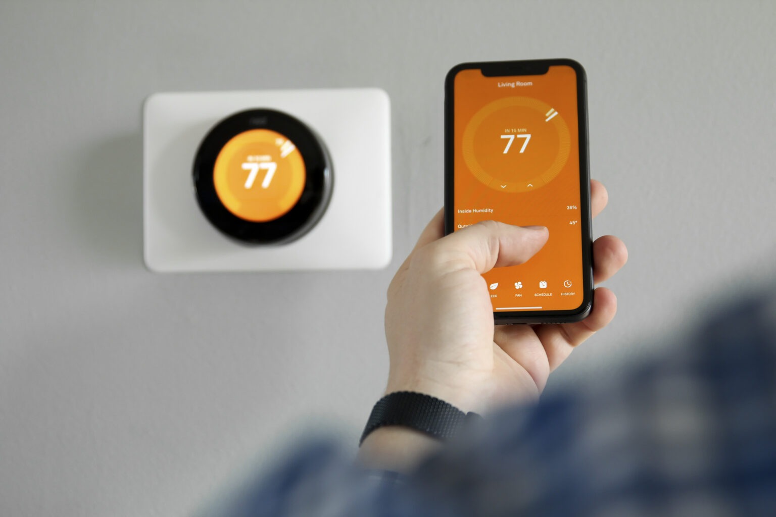 A smart thermostat is shown next to a phone mirroring its display and being operated by a user.