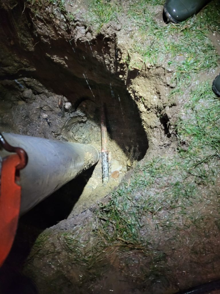 A muddy hole in the ground reveals a leaking pipe, emphasizing the need for emergency plumbing services. 