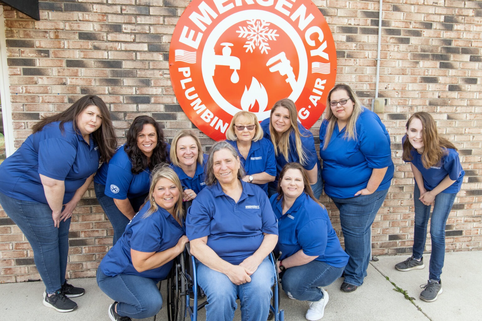 A group of women, wearing matching blue shirts and jeans, poses in front of an Emergency Plumbing sign.