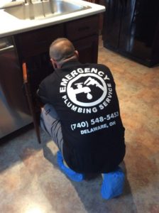 A plumber, with a black shirt displaying "Emergency Plumbing Service (740) 548-5453 Delaware, OH", kneels and works under a kitchen sink