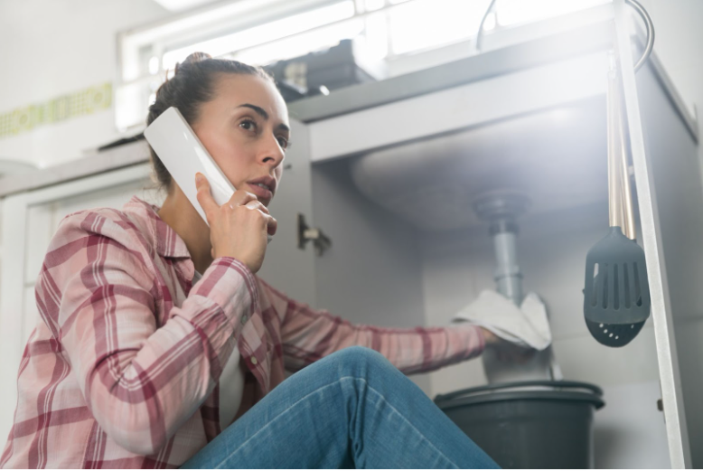 Woman is on the phone with a plumber while inspecting a leaky pipe under her sink