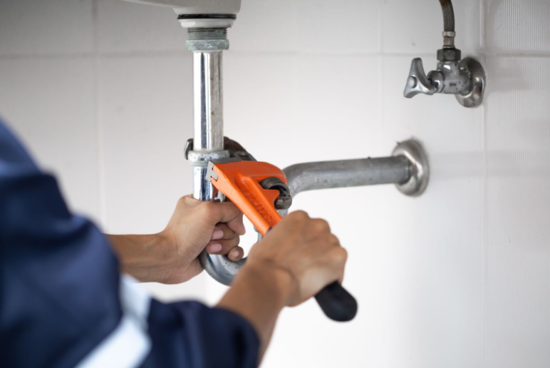 A plumber uses an orange wrench to repair a pipe under a sink