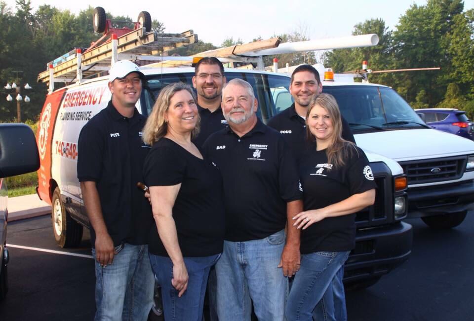 A group of six people stand smiling in front of an "Emergency Plumbing Service" van