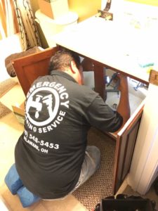 A plumber kneels to repair the plumbing under a bathroom sink.